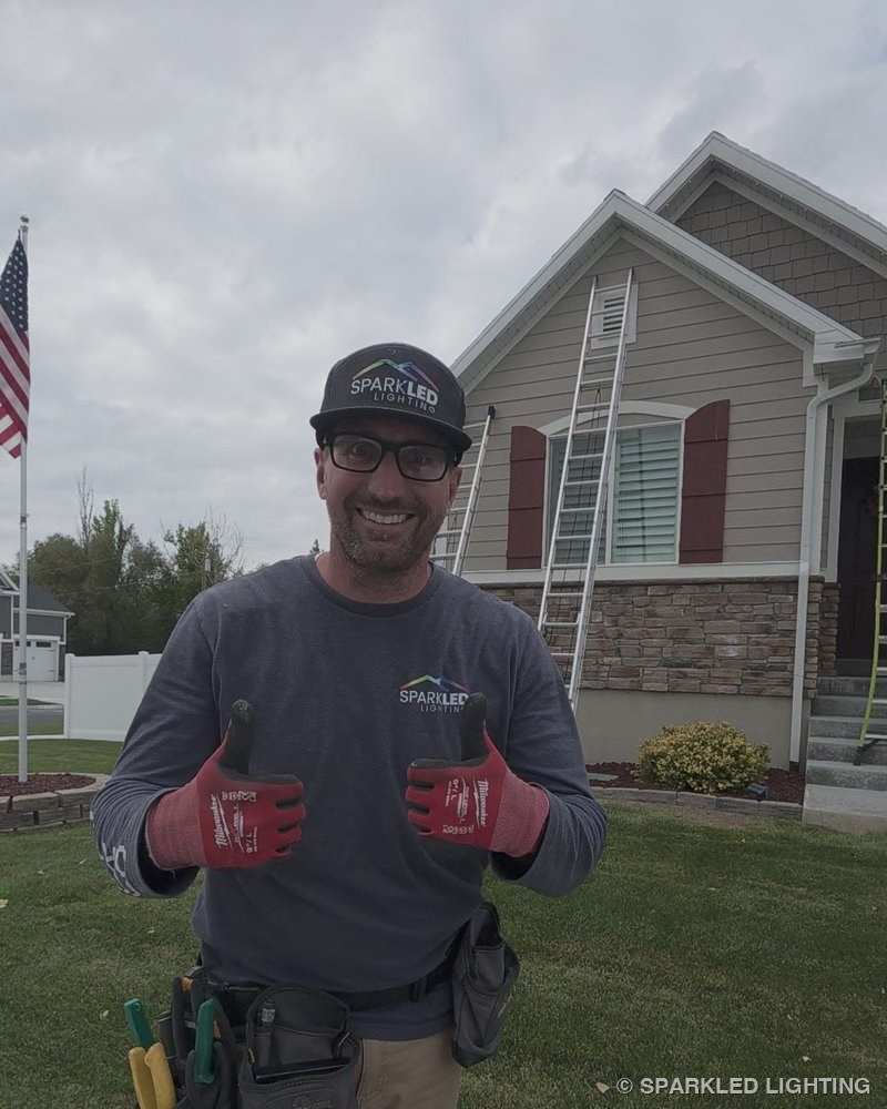Ryan Kennedy, founder of Sparkled Lighting, in a Sparkled cap and tool belt giving two thumbs up on an install day in front of a Utah home