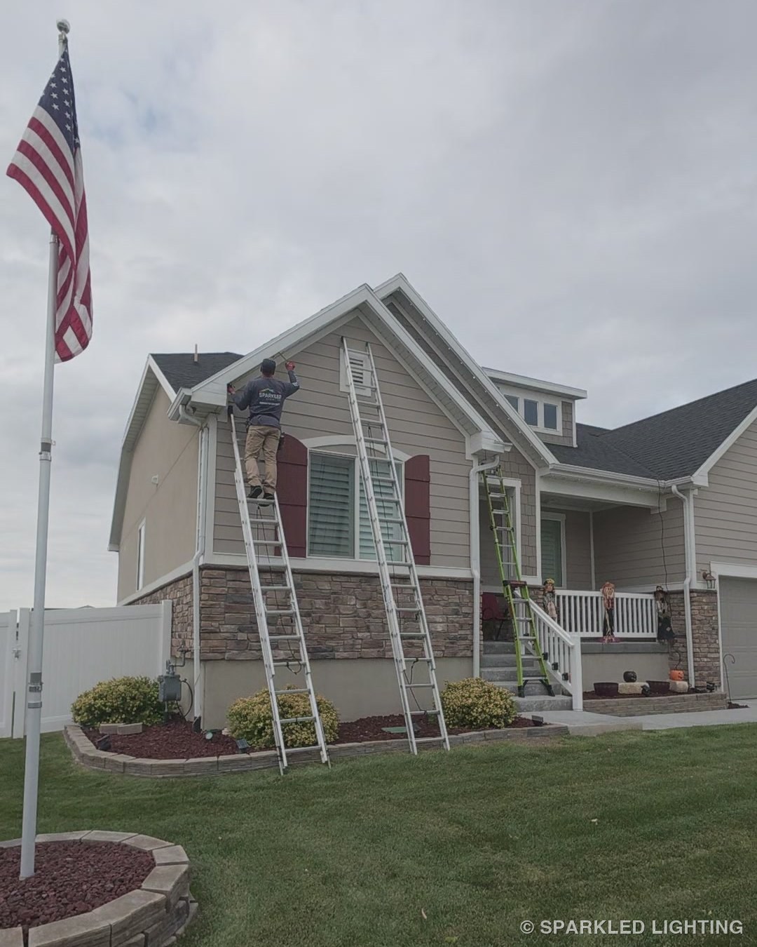 Ryan of Sparkled Lighting on a ladder installing permanent LED lights at the gable of a Utah home, American flag in the yard